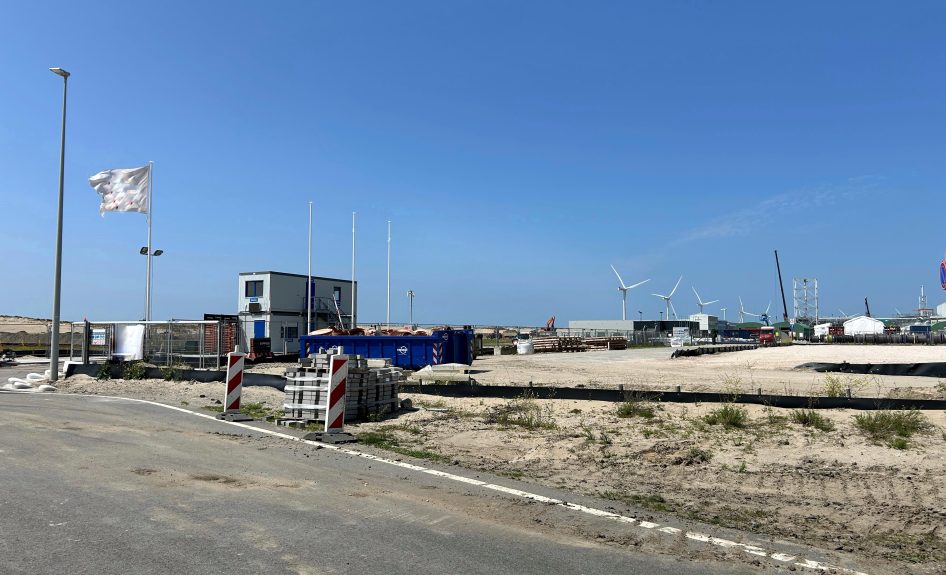 Bouwlocatie toekomstige waterstoffabriek op de Maasvlakte, met windmolens op de achtergrond en eerste voorbereidende werkzaamheden in beeld, waaronder containers, bouwmaterialen en tijdelijke kantoorunits.